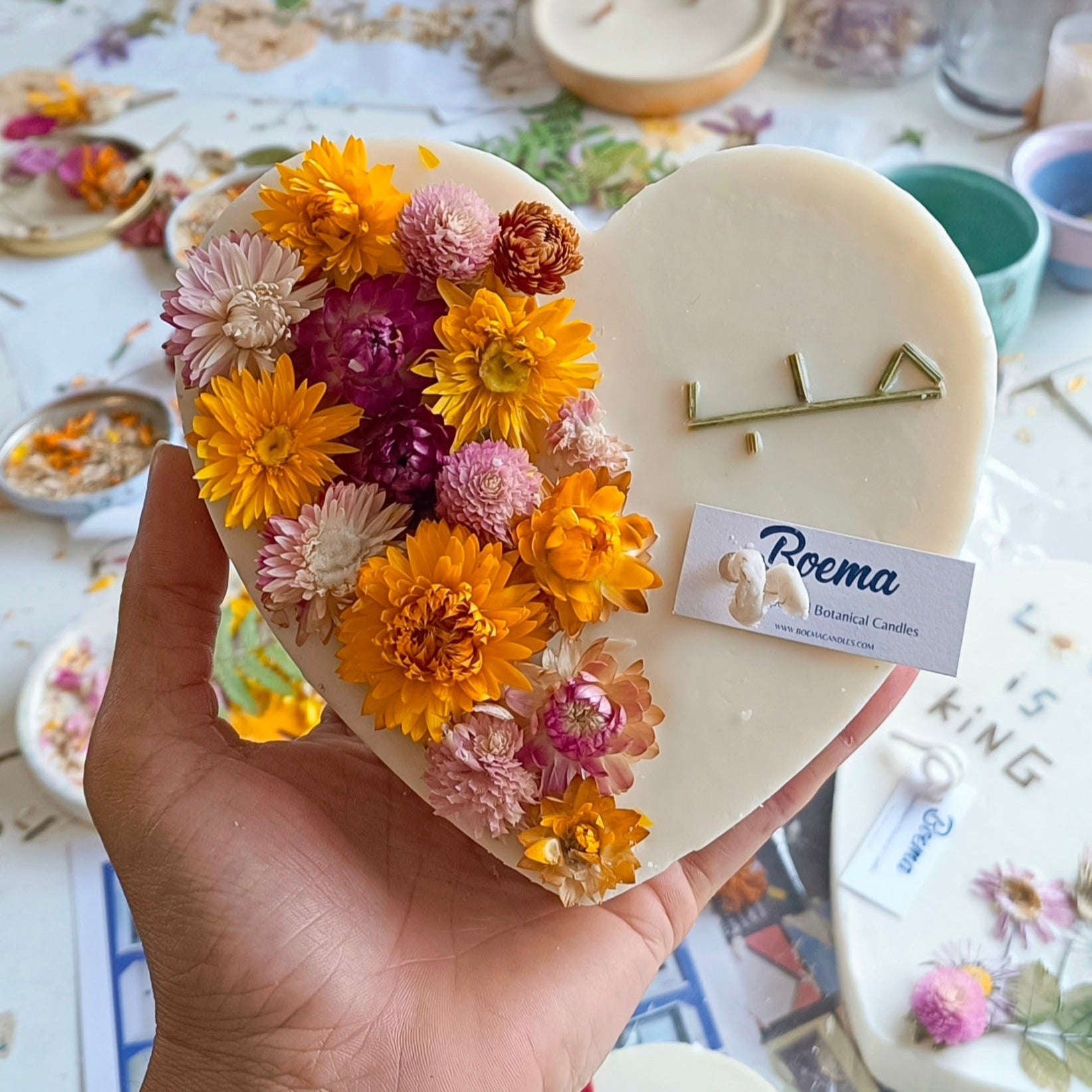 Heart-shaped floral arrangement with 'Poema' branding held by a hand on a decorative table.