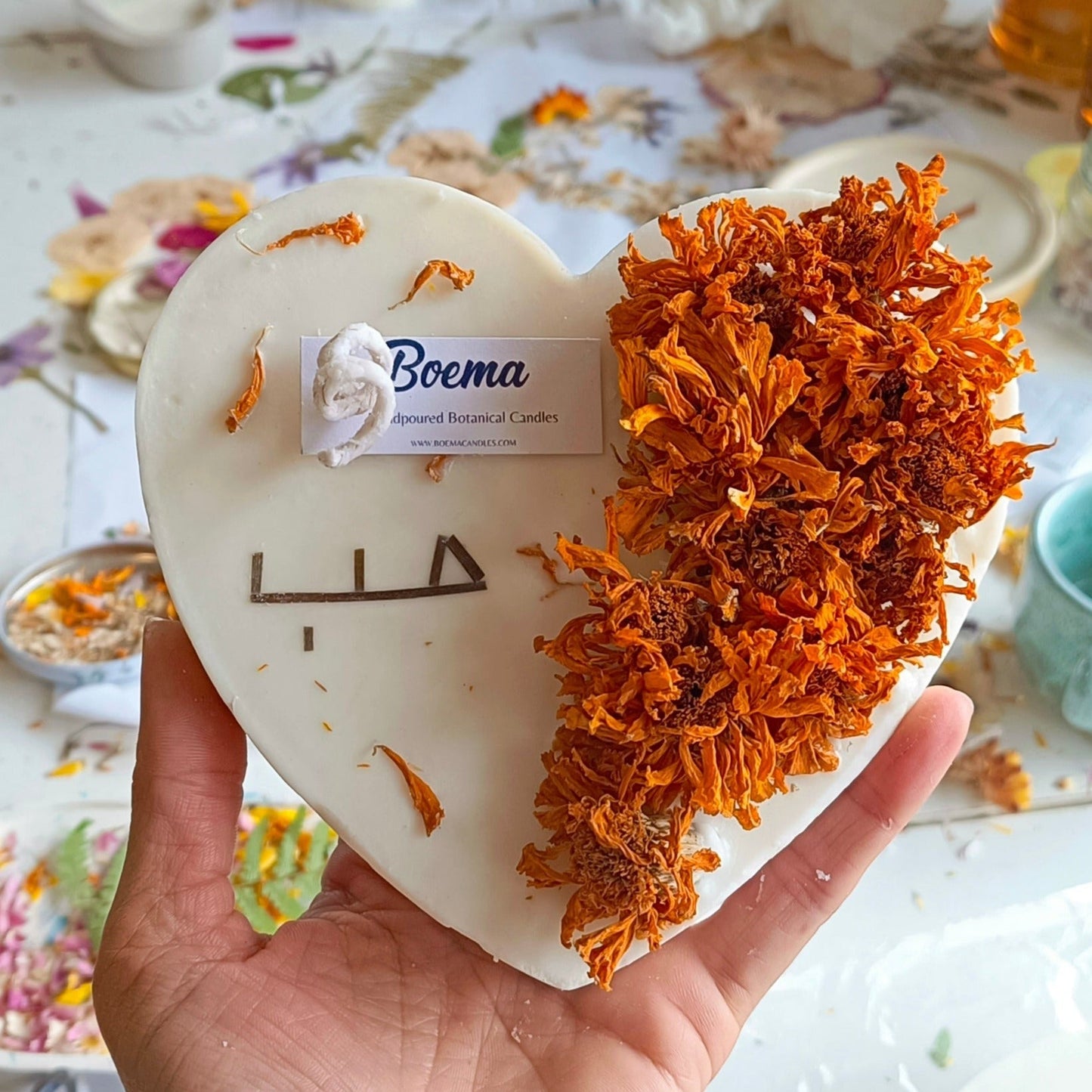 A hand holding a large heart-shaped candle decorated with dried flowers, with the brand Boema visible on the candle.